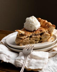 slice of apple pie with scoop of vanilla icecream on top, on a stack of plates with a fork and on a white napkin