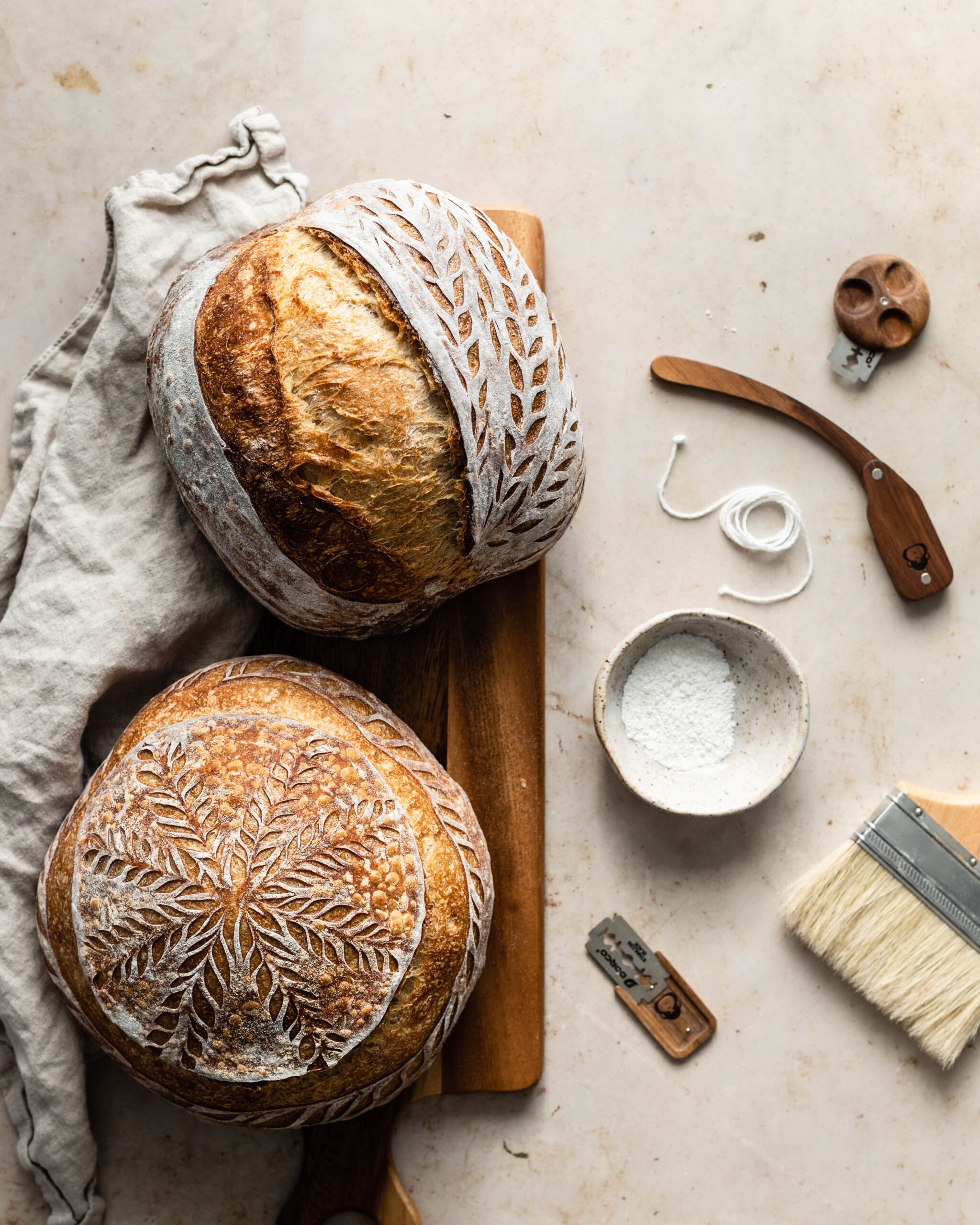 Two sourdough loaves on a cutting board with bread scoring tools