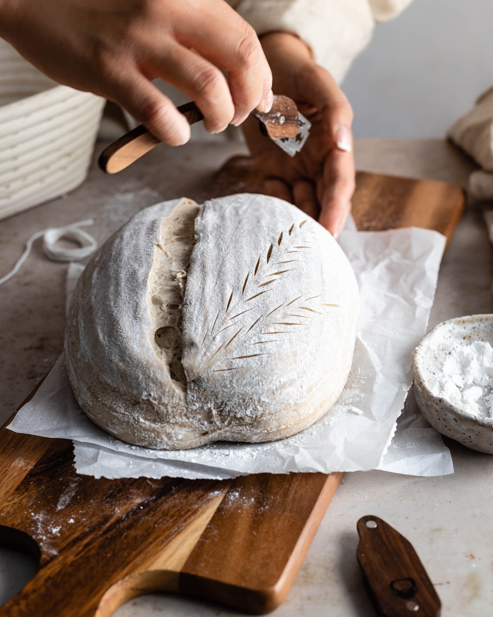 Hands holding a lame creating a wheat stalk pattern on dough