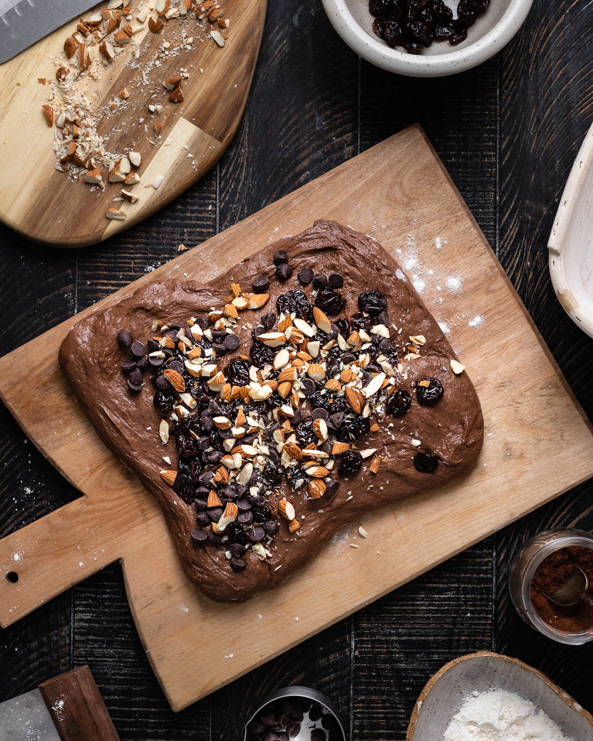 chocolate dough on wooden cutting board with cherries, almonds and chocolate chips on top