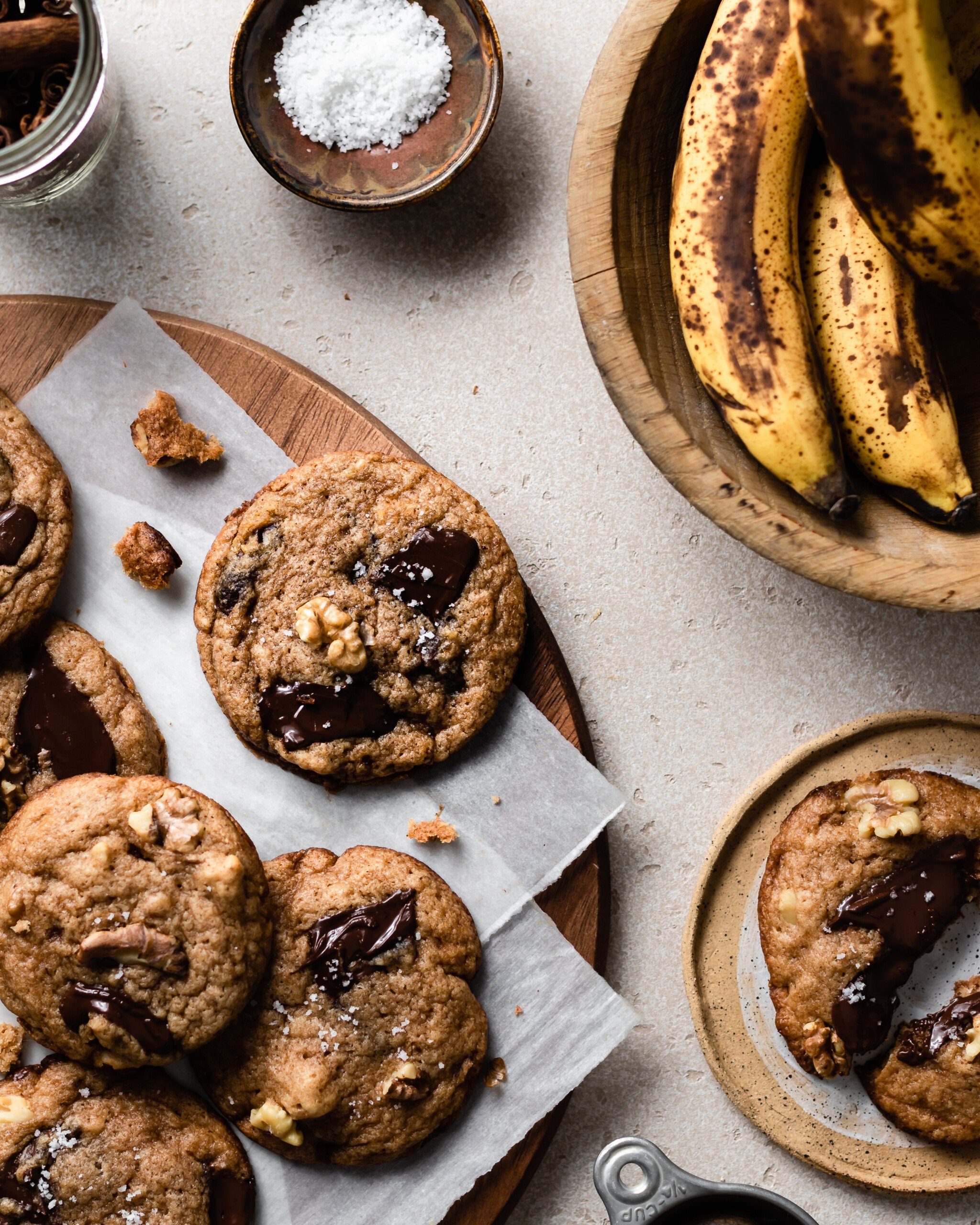 Chocolate cookies with melted chocolate pool and walnuts on a cutting board lined with parchment paper with wood bowl containing bananas and a cookie cut in half. 