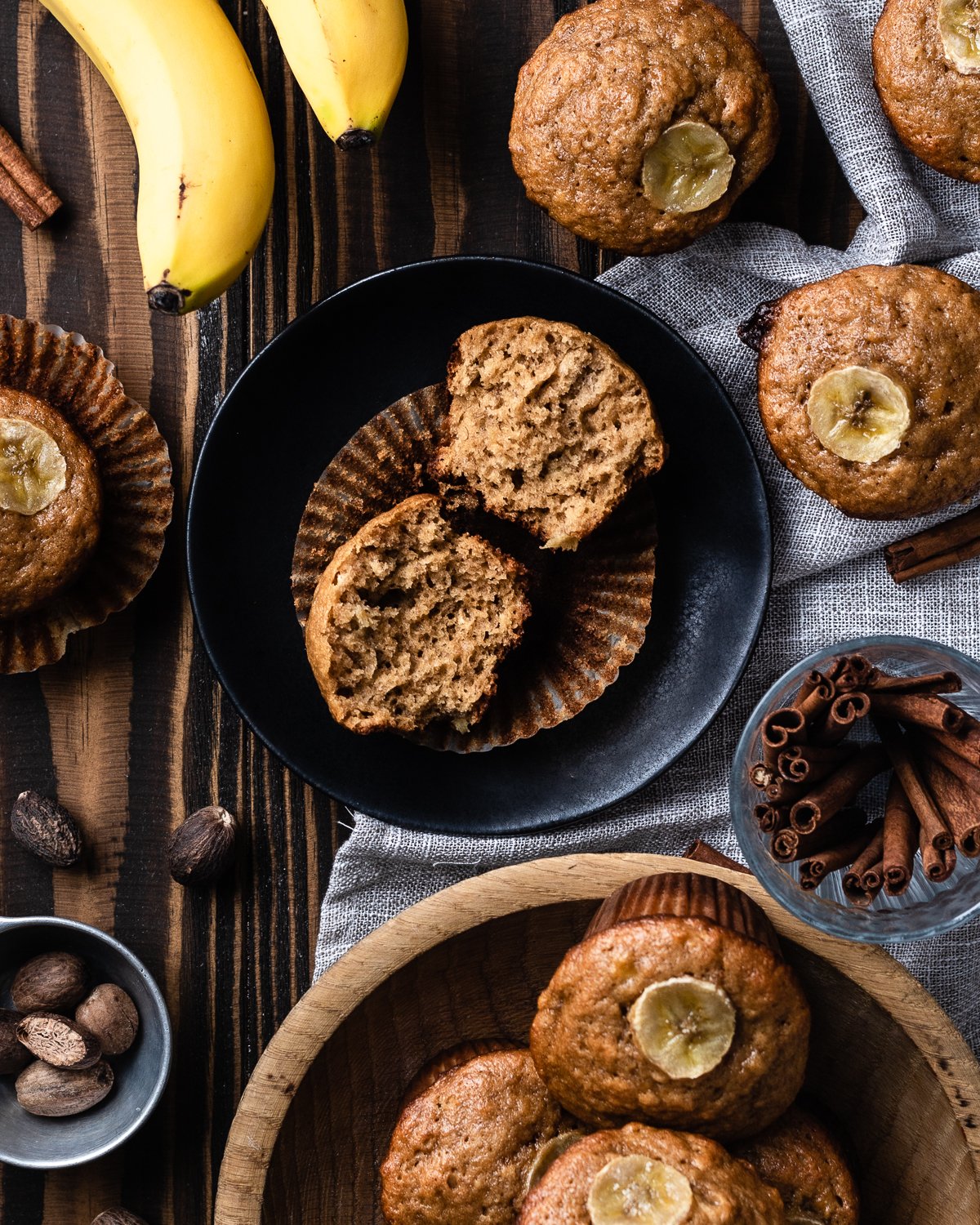 banana muffin cut in half showing plush crumb on a muffin liner on top of a black plate with muffins on a table