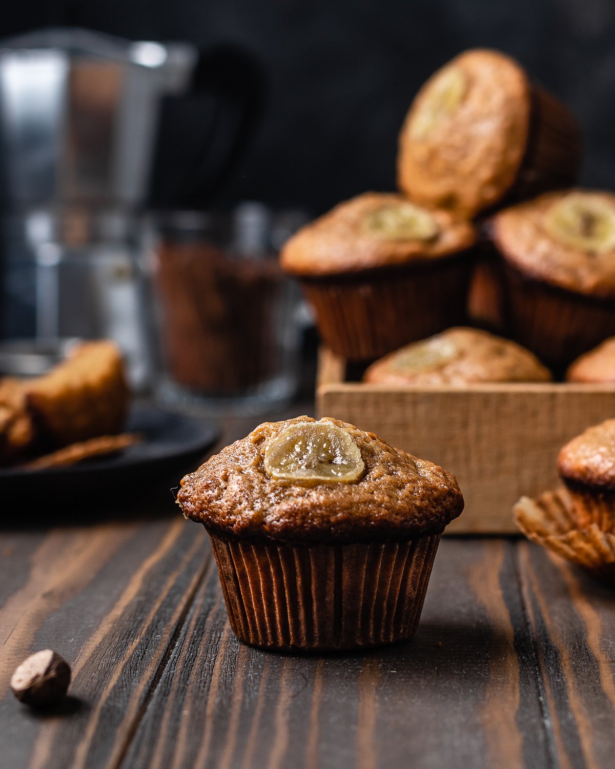 banana muffin with a slice of banana on top and bananas in a pie inside a woodbox in the background