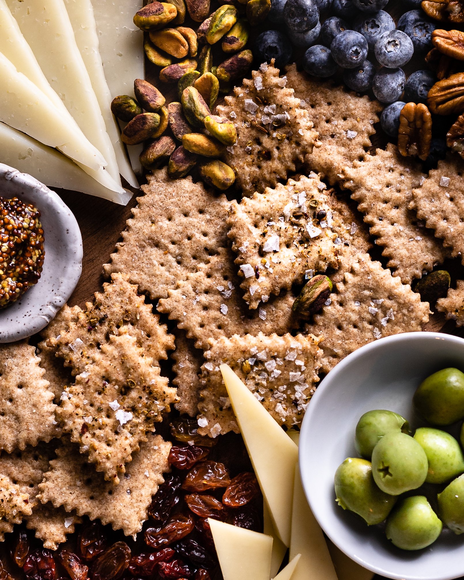 sourdough crackers in the center of a cheese board with cheeses, raisins, olives and mustard