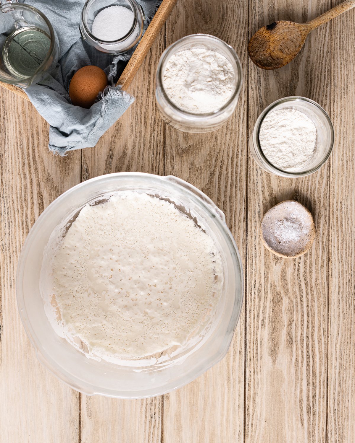 bubbly sponge in a large mixing bowl with wooden spoon, flours in jars, salt, egg, sugar and oil