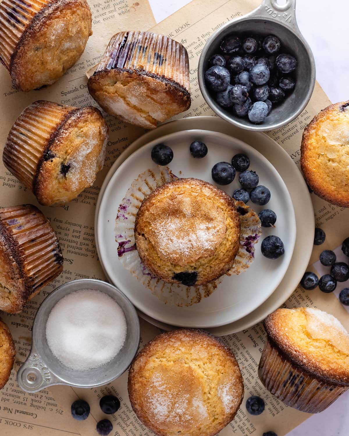 Blueberry muffin with muffin liner peeled off on top of a plate over a few pages of books surrounded by a cup of blueberries, sugar and blueberry muffins.