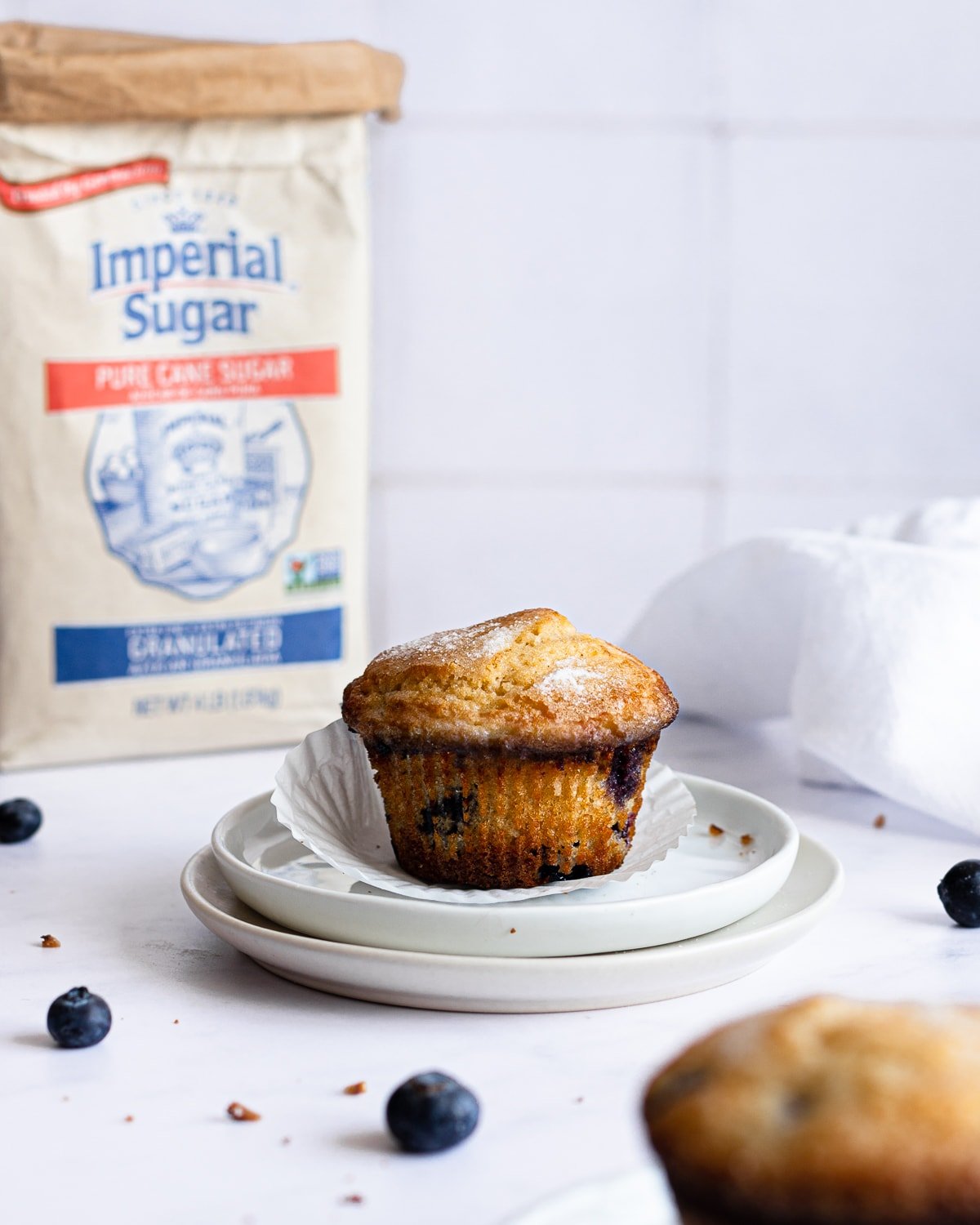 Blueberry muffin on a stack of two small plates with a bag of sugar and white napkin in the background.