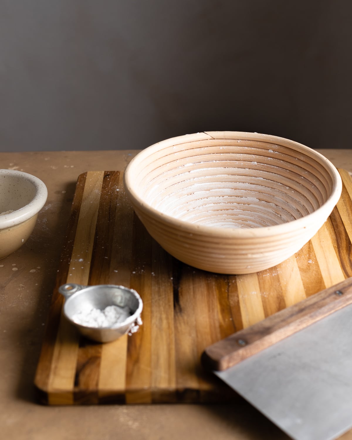 round wood banneton on a wooden cutting board with a bench knife and a metal cup with rice flour