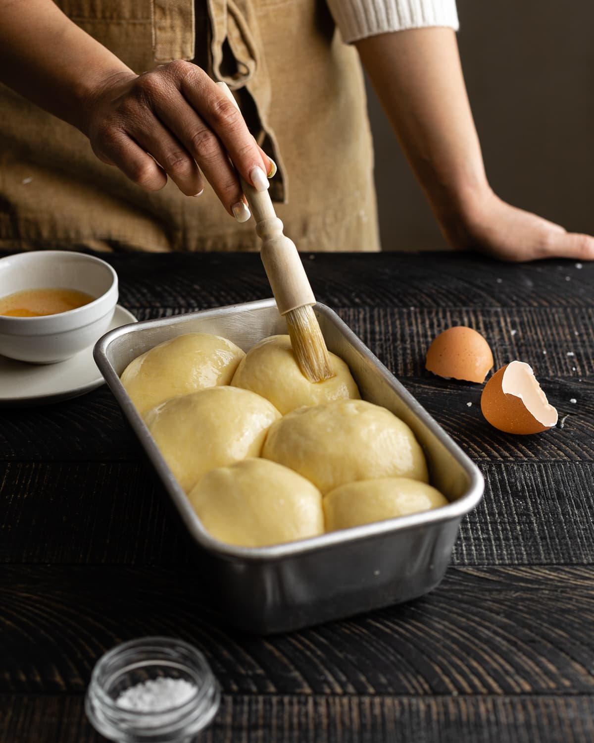 Hand holding pastry brush brushing egg wash on dough inside loaf pan