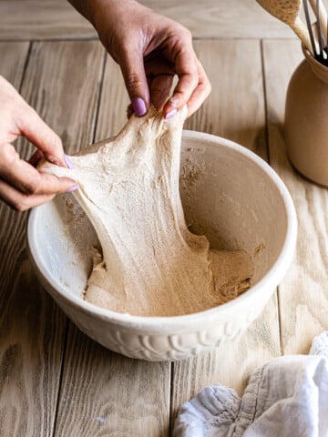 Hands stretching out piece of dough in a white mixing bowl