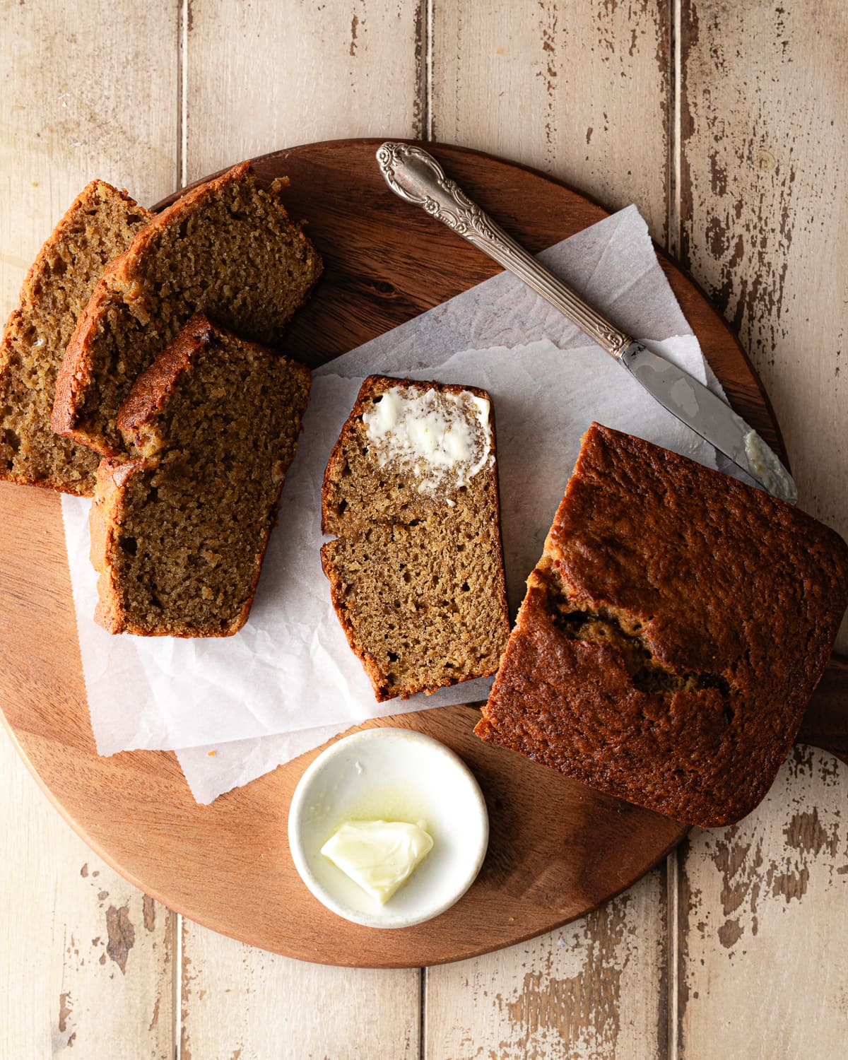 Sliced banana bread on a round cutting board and parchment paper with a pat of butter in small plate