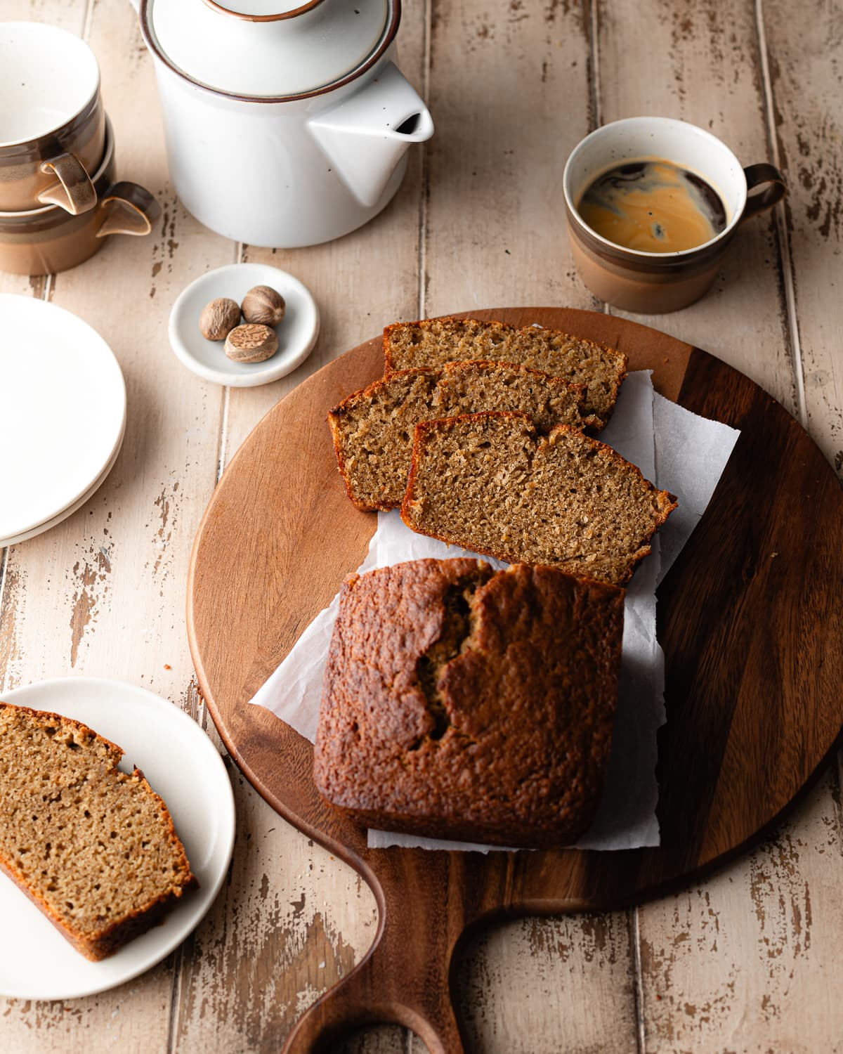 Sliced banana bread on round cutting board with nutmeg, coffee and a coffee pot