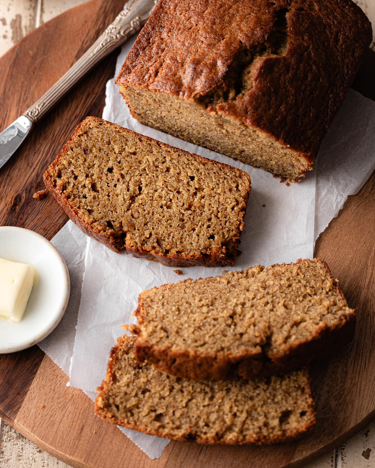 Sliced banana bread on a round cutting board and parchment paper with a pat of butter in small plate