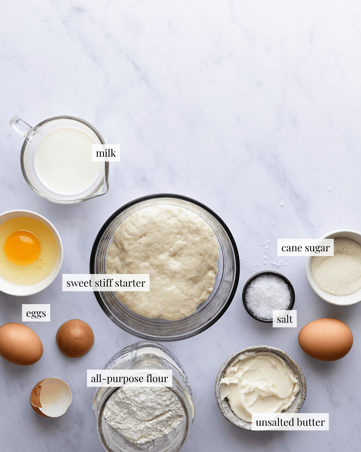 ingredients for sourdough rolls, milk in a small pitcher, egg in a bowl, starter in a glass bowl, all-purpose flour in a large jar, butter in a small bowl, salt in a small bowl and sugar in a small bowl. 