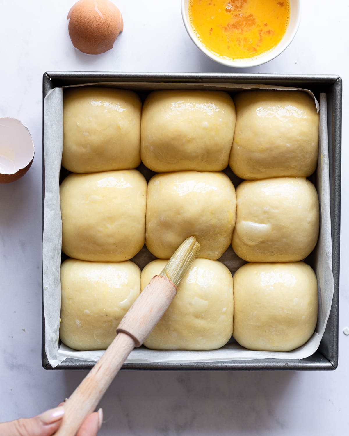 hand brushing rolls with egg wash in a metal baking pan. 
