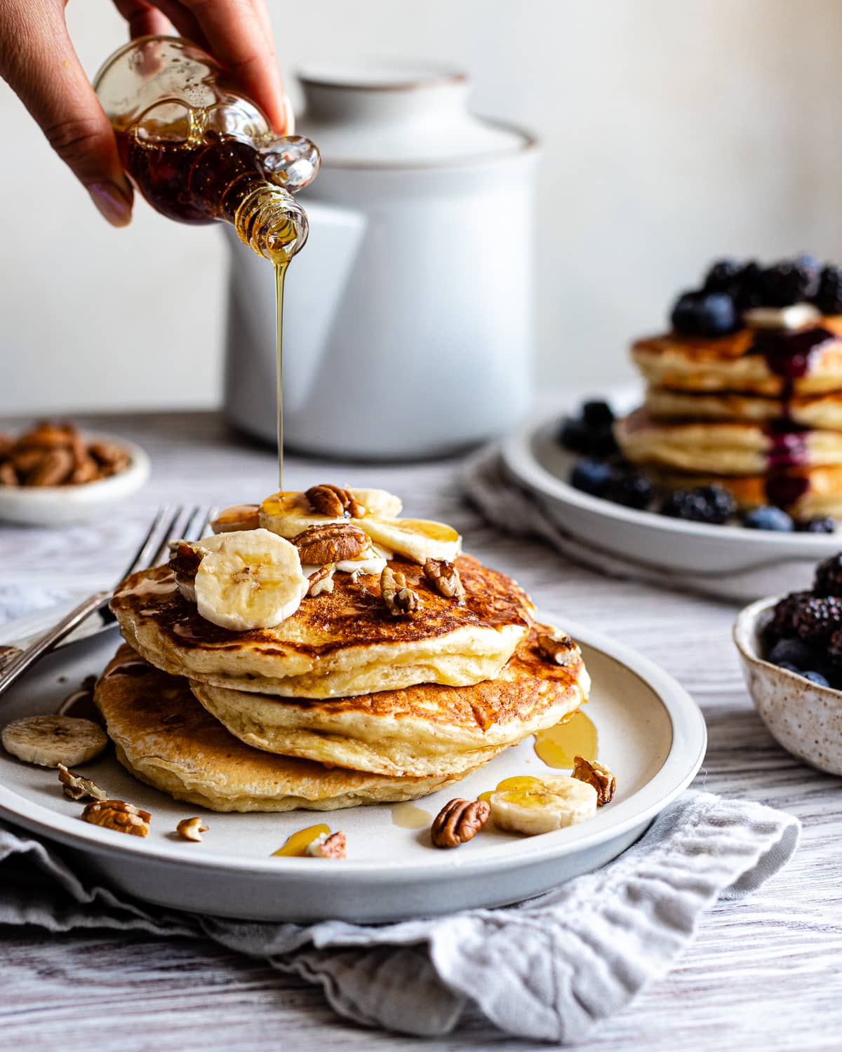 Hand pouring maple syrup on stack of 3 pancakes topped with bananas and pecans.