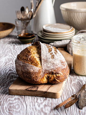 Sourdough bread on a wooden cutting board beside a bubbly sourdough starter in a mason jar.
