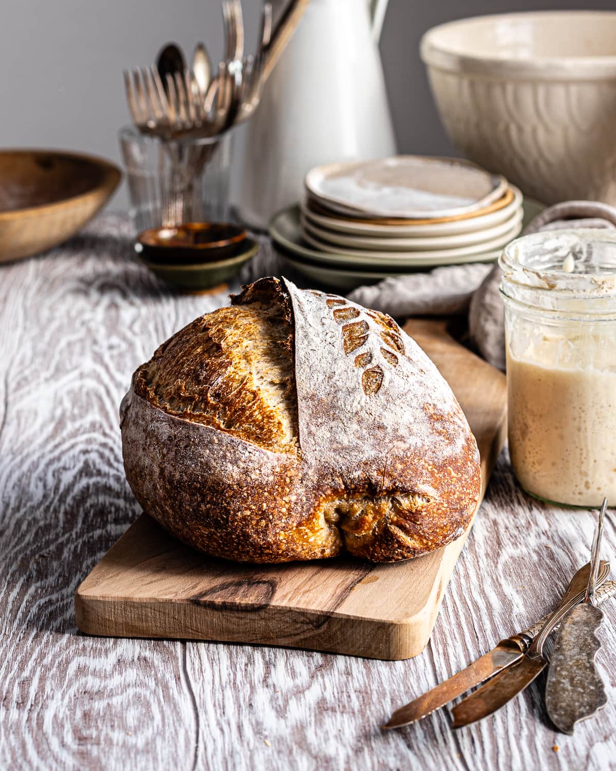 Sourdough bread on a wooden cutting board beside a bubbly sourdough starter in a mason jar.