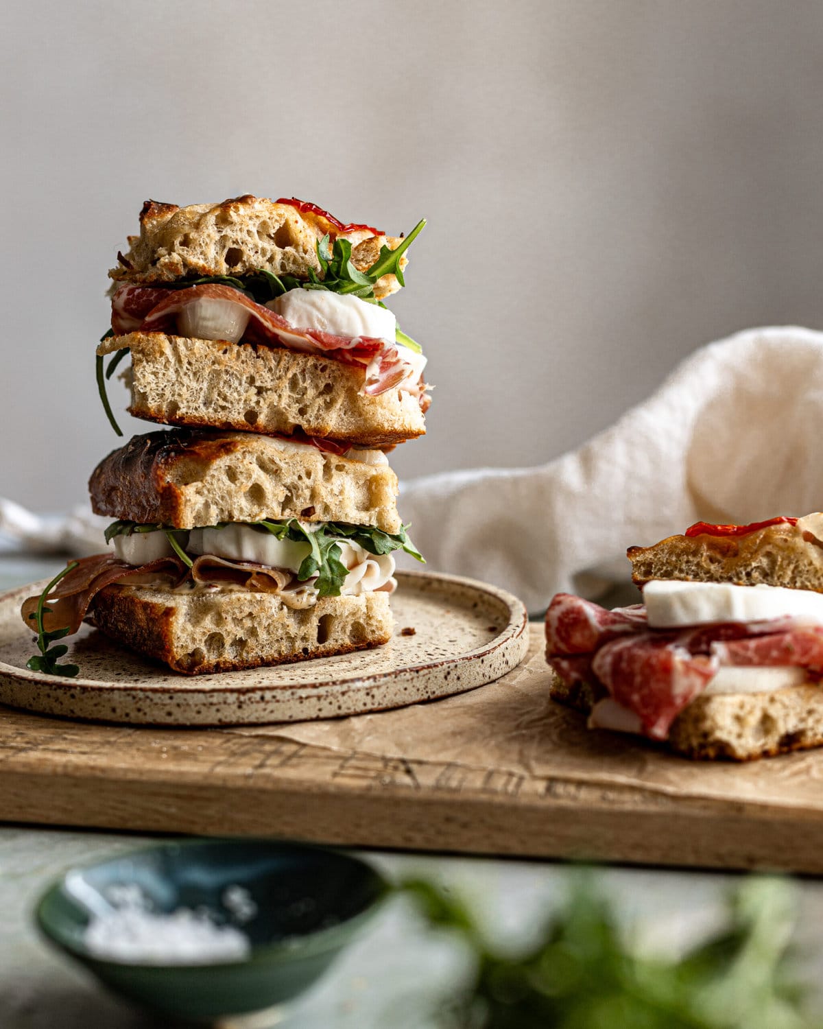 A stack of focaccia sandwiches with prosciutto, arugula and mozzarella cheese on a plate. 