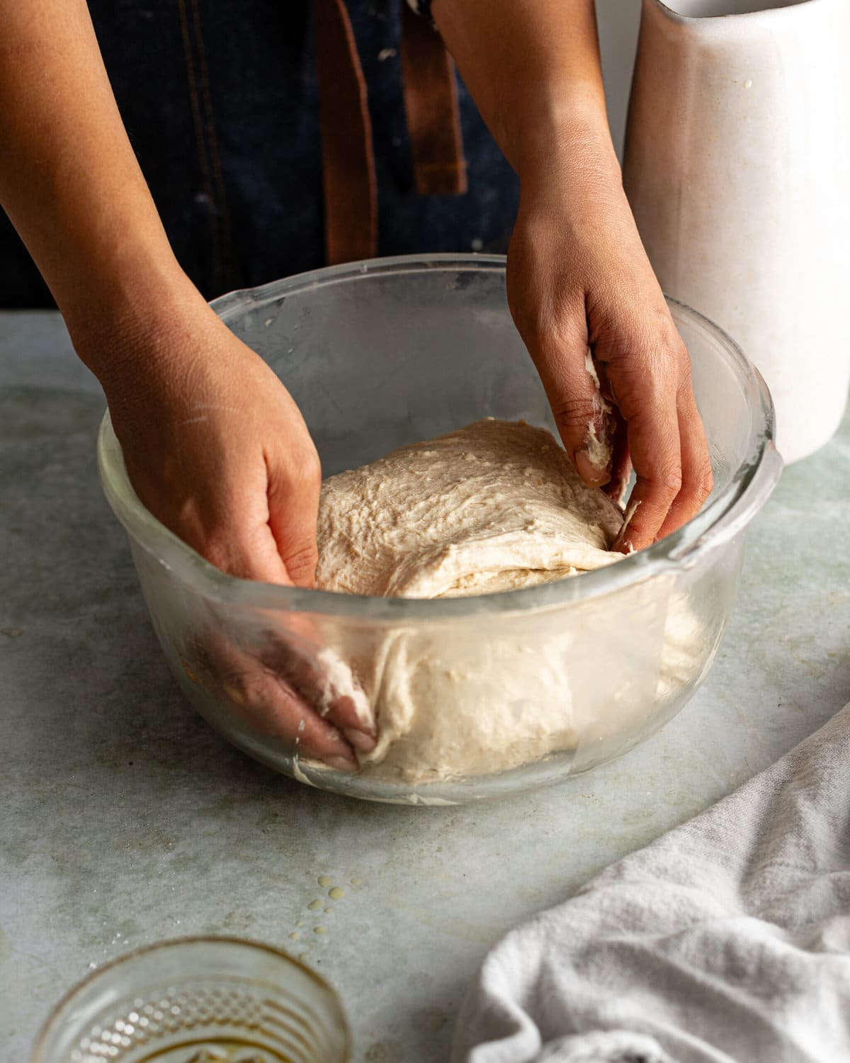 Hands folding a piece of dough inside a glass mixing bowl.