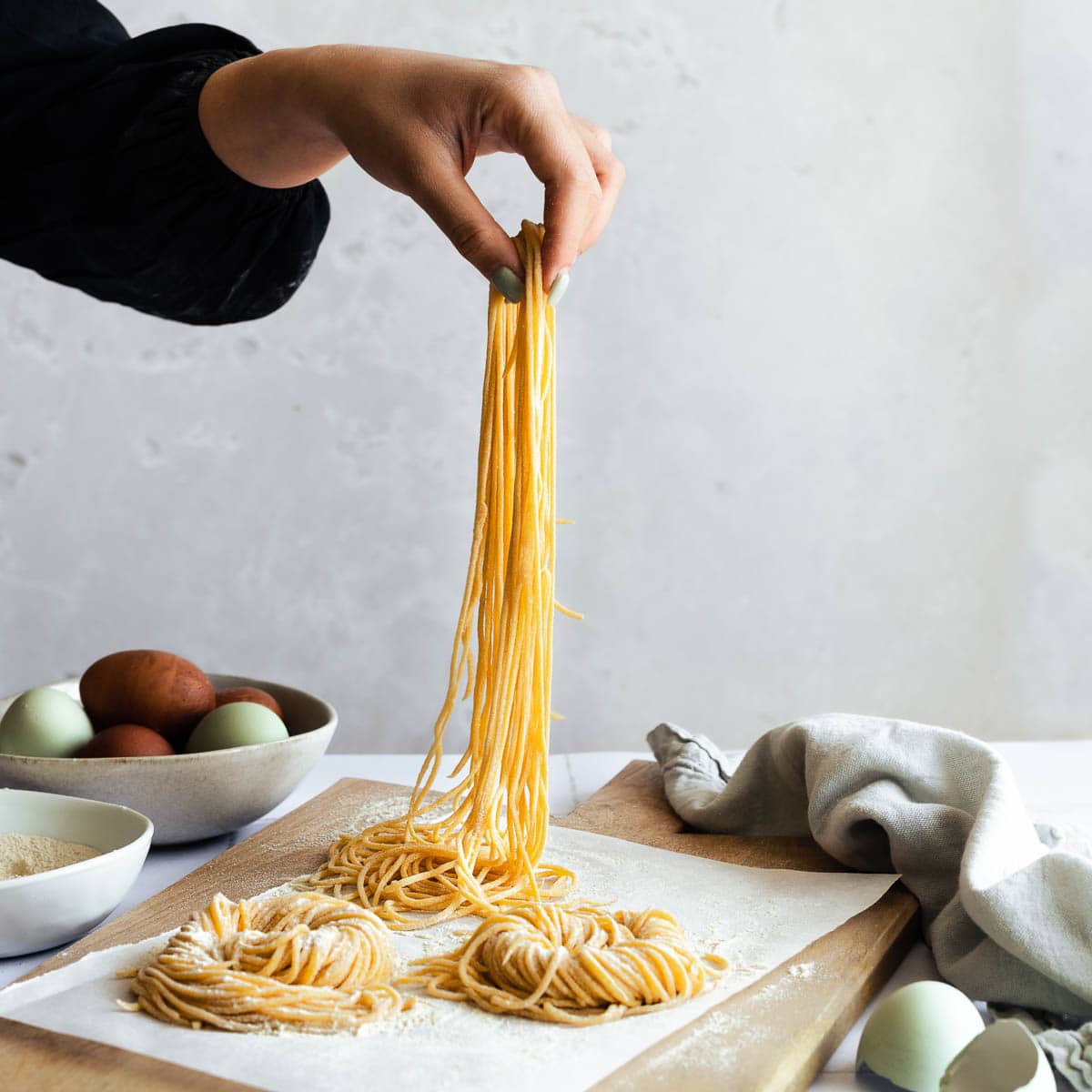 Hand holding up uncooked pasta with nests of pasta on a cutting board. 