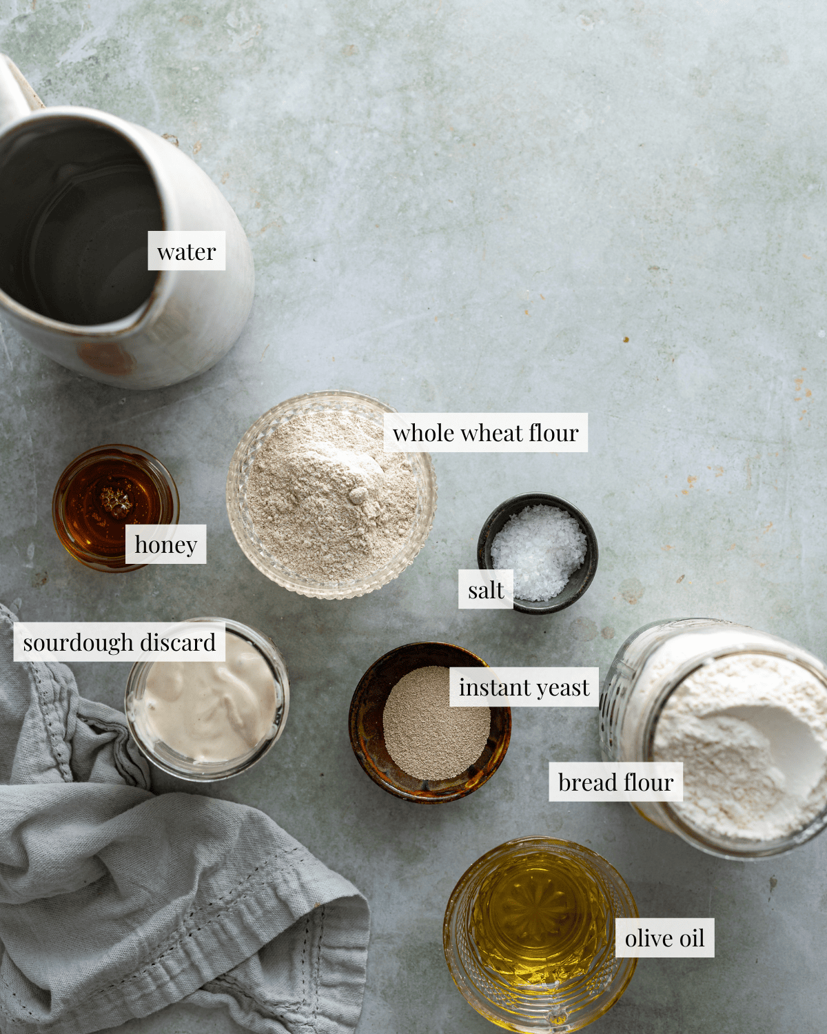 Ingredients for a focaccia, water in a pitcher, flour in jars, and yeast, honey, and salt in a small bowls and olive oil in a glass cup.