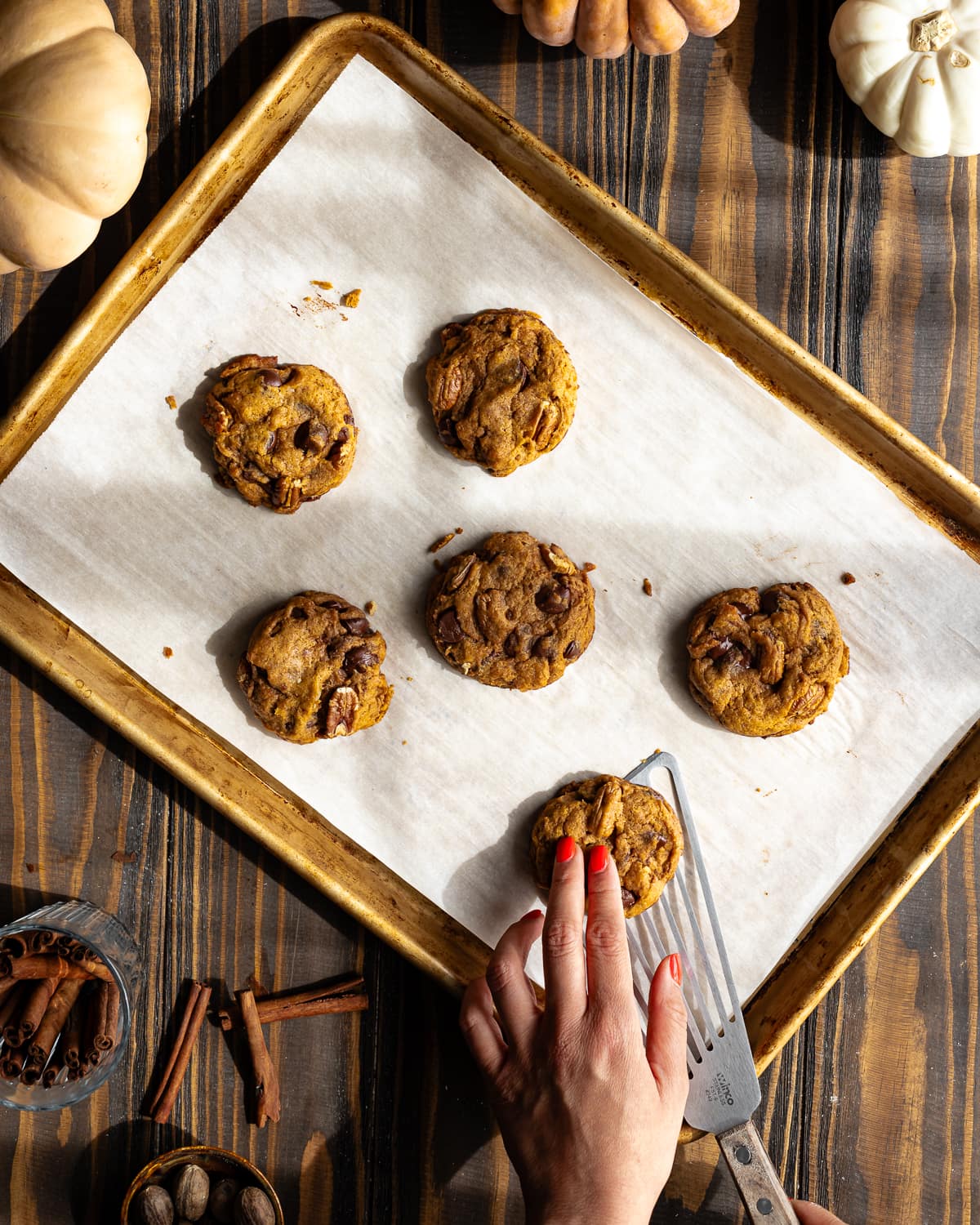Pumpkin cookies on a parchment lined baking sheet with a hand scooping up a cookie with a fish spatula. 