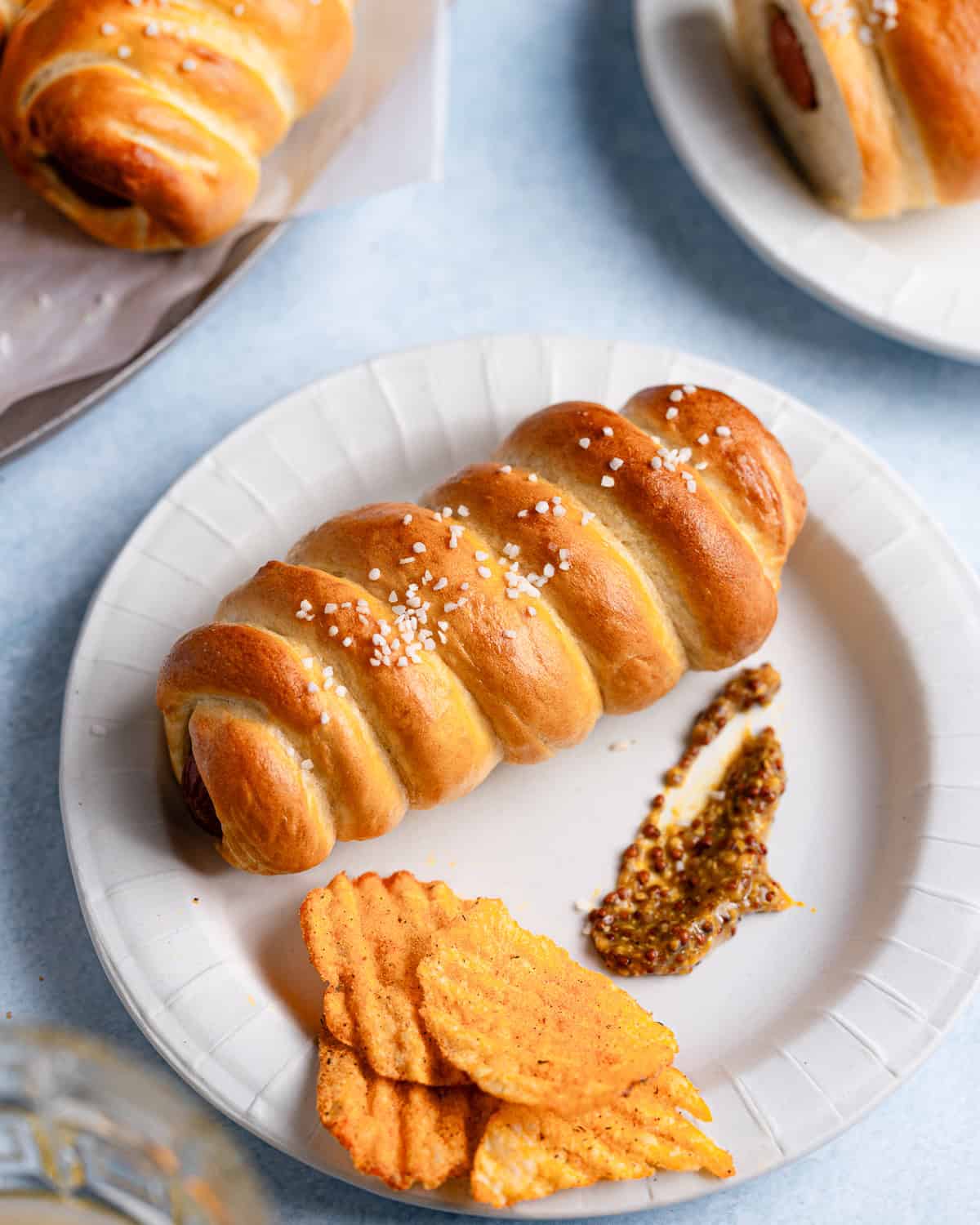 Pretzel dog on a small plate with chips and mustard. 