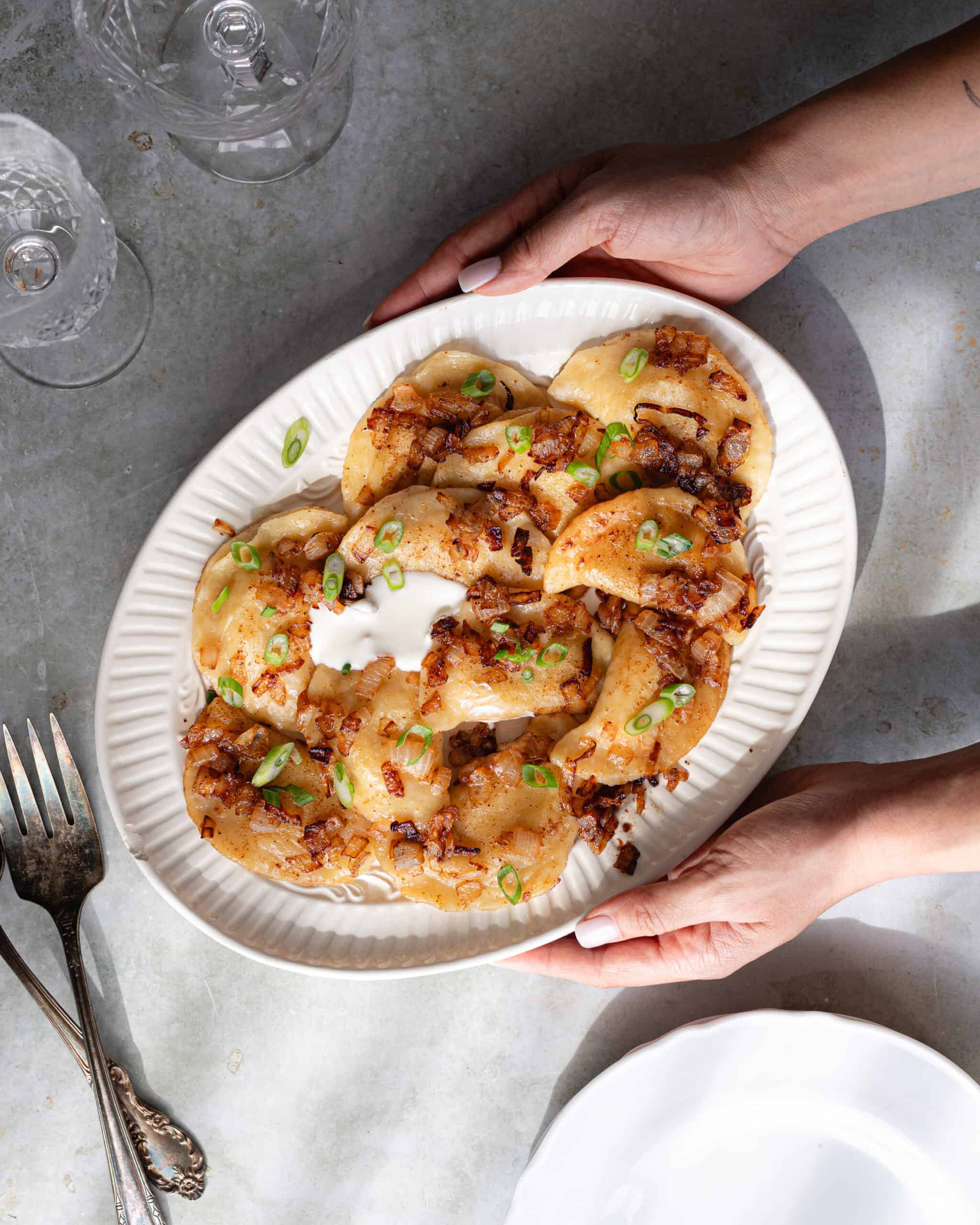 Hands holding a white platter of pierogi topped with sour cream, caramelized onions and scallions.