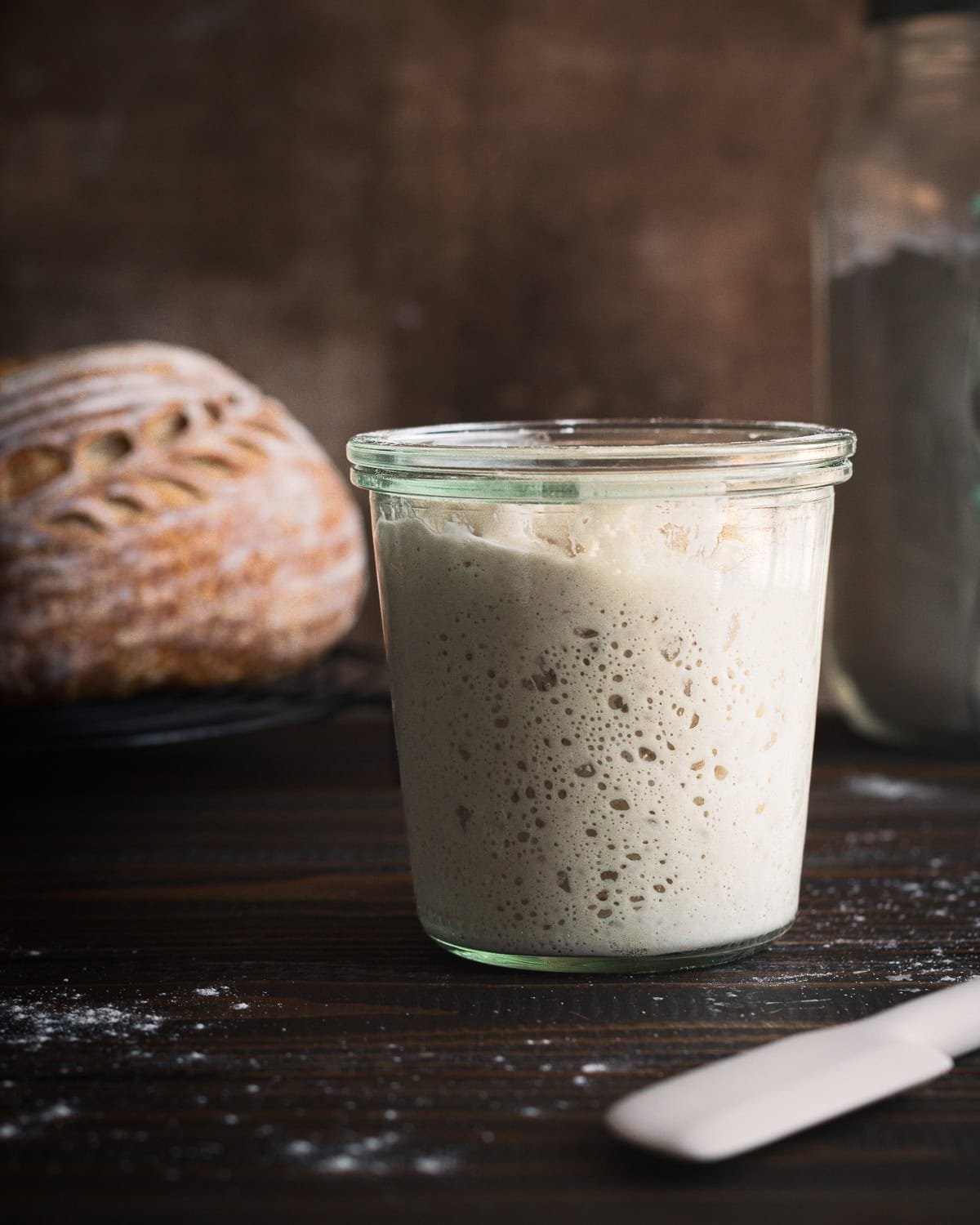 Sourdough starter in a jar with bread in the background.